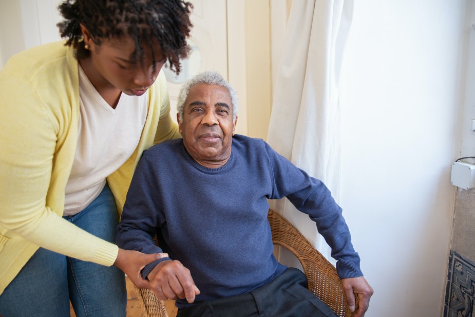 A black woman carer helps an elder from his chair